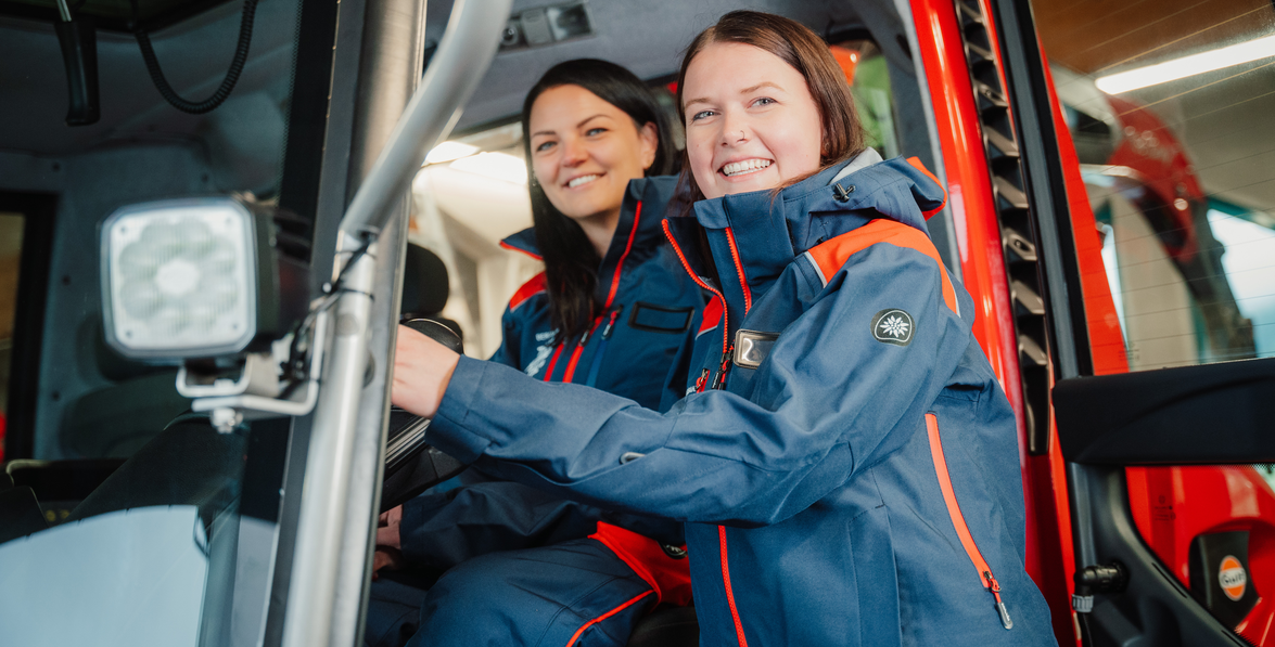 Zwei Frauen in Arbeitskleidung lächeln in einer Pistenraupe