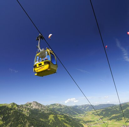 Gelbe Seilbahn vor blauem Himmel und grüner Berglandschaft mit Gleitschirmfliegern