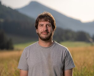 Mann mit Bart und grauem T-Shirt vor Landschaft mit Berg