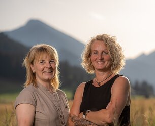 Zwei Frauen stehen lächelnd vor Bergkulisse im Feld