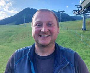 Lächelnder Mann mit blauer Weste vor grüner Wiese und Bergkulisse mit Seilbahn