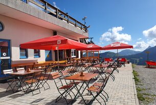Terrasse mit Tischen, Stühlen und roten Sonnenschirmen vor einem Gebäude unter blauem Himmel