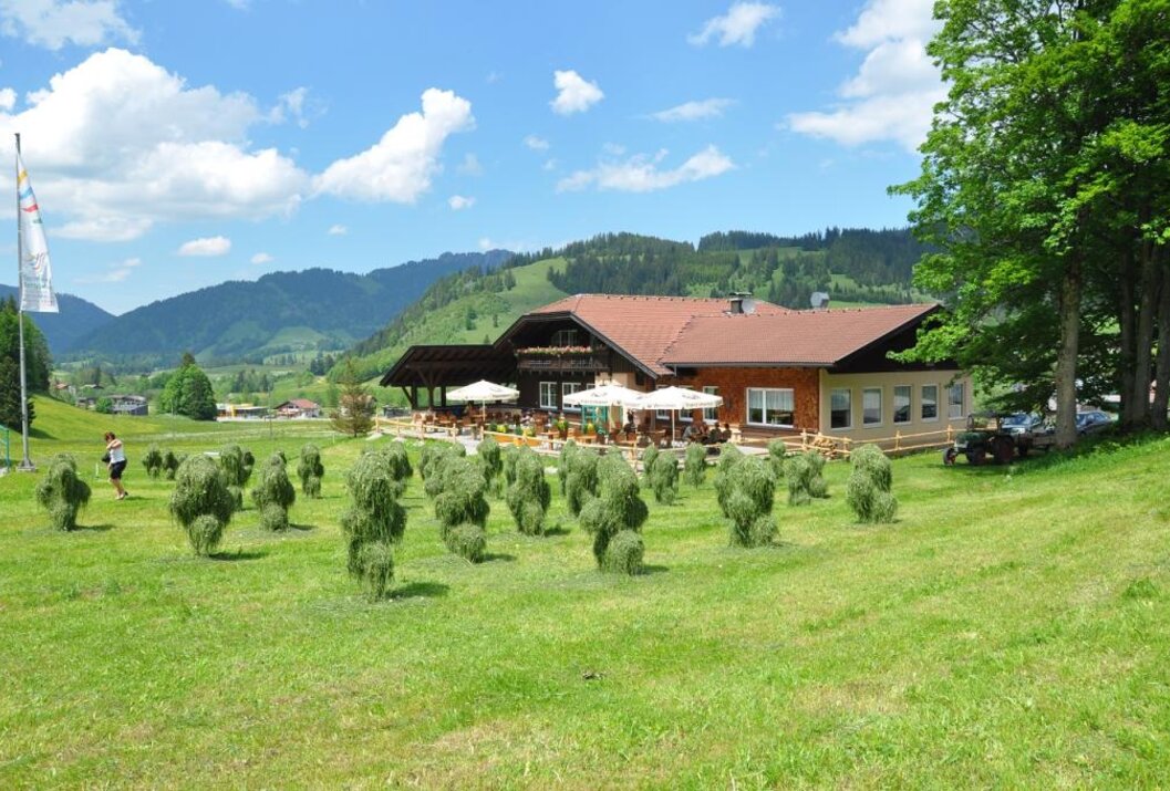 Landhaus mit Terrasse auf grüner Wiese, im Hintergrund Berge und blauer Himmel mit Wolken